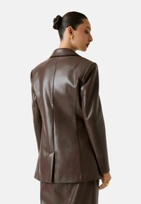 Blazer in pelle marrone con design aderente, revers affilati e un'unica spaccatura posteriore. La texture lucida ne accentua l'aspetto elegante.