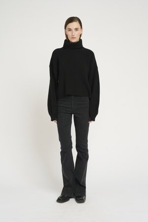 Svart ribbet turtleneck-genser med vide ermer, kombinert med høyt midjade sorte bootcut-jeans og sorte sko, satt mot en hvit bakgrunn.