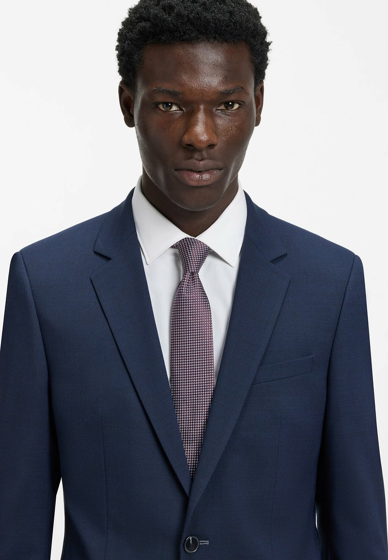 Veste de costume bleu marine à revers cranté et fermeture à un bouton, associée à une chemise blanche et une cravate violette texturée avec un motif pied-de-poule.