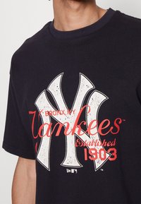 Svart bomullströja med en stor vit Yankees-logotyp, med röd text som säger "BRONX, NY" och "Etabl. 1903." Rund halsringning, korta ärmar.