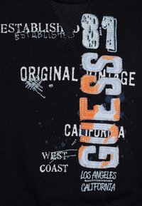Tela negra con texto y gráficos bordados en blanco, azul y naranja. Presenta "GUESS" y "Establecido en 1981" de manera destacada.