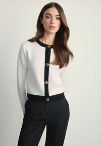 Biały kardigan z czarnymi wykończeniami i złotymi guzikami, z okrągłym dekoltem i ściągaczami, w komplecie z czarnymi spodniami.