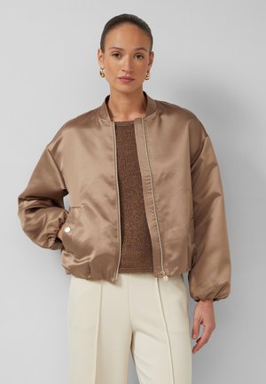 Femme aux cheveux plaqués en arrière portant des boucles d'oreilles en or, une veste bomber en satin marron clair, un haut en maille marron et un pantalon plissé crème, debout devant un mur gris.