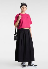 Růžové zkrácené tričko, černá maxi sukně, černá čepice s bílým potiskem, černá crossbody taška a černé tenisky s bílými akcenty.