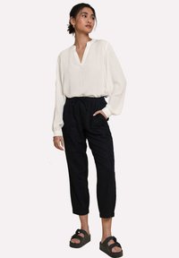 Bílá dlouhá blůza s výstřihem do V a elastickými manžetami, spárovaná s černými zúženými kalhotami s elastickým pasem a kapsami. Černé sandály.