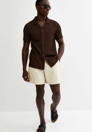 Mann trägt dunkelbraunes, kurzärmeliges Strickhemd, cremefarbene Shorts, schwarze Sandalen und Sonnenbrille, geht mit einer Hand in der Hosentasche.