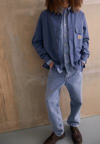 Blauwe button-up shirt over een lichtblauw denim shirt, gecombineerd met vervaagde blauwe jeans en donkerbruine schoenen, met zak op het shirt.