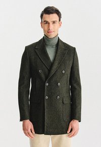 Dwurzędowy zielony wełniany płaszcz z dużymi guzikami i dwoma kieszeniami z przodu, noszony na dopasowanym jasnooliwkowym golfie.