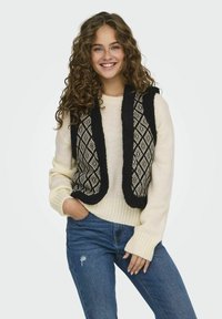 Gebreide trui in crème met geribde mouwen, gecombineerd met een zwart en crème vest met een patroon van ruitvormen, gedragen over een blauwe jeans.