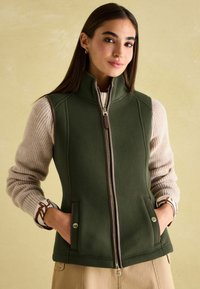 Grüne Fleece-Weste mit hohem Kragen, brazip und Taschen. Über einem beigefarbenen Rippensweater getragen. Verfügt über goldene Druckknopfakzente.