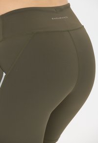 Läheltä kuvattu oliivinvihreä korkeavyötäröinen urheilulegginsit, joissa on näkyvät saumat ja "ENDURANCE"-logo vyötäröllä.