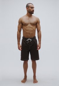 Hnedé plavkové šortky s bielym šnúrkou, s elastickým pásom a bočnými detailmi. Model stojí bosý na neutrálom pozadí.