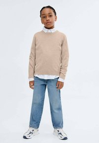 Beige neulottu villapaita, jossa on pitkät hihat, valkoisen kauluspuseron päällä, yhdistettynä vaaleansinsisiin löysiin farkkuihin ja valkoisiin tennareihin.