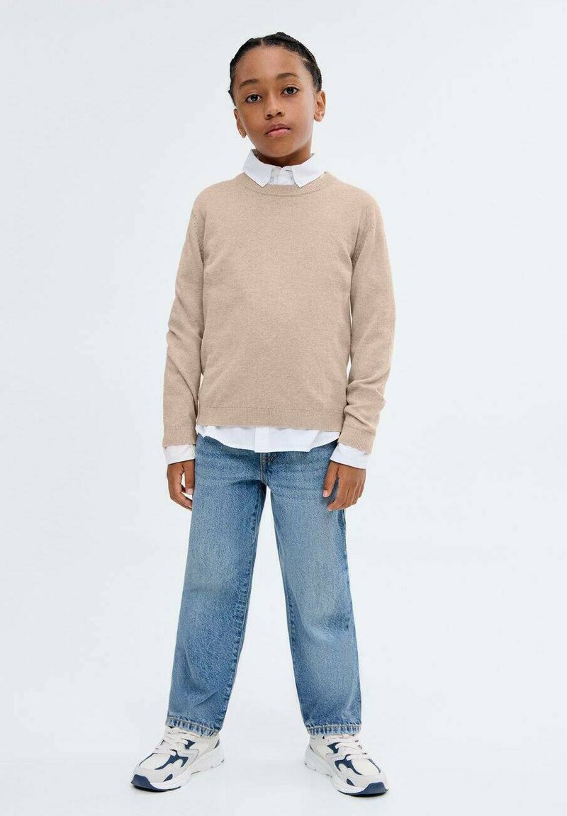 Béžový pletený svetr s dlouhými rukávy, přes bílou košili s límečkem, spárovaný se světlými modrými volnými džíny a bílými teniskami.