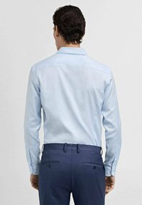 Camisa de vestir azul claro con una textura suave, cuello puntiagudo y puños abotonados, combinada con pantalones oscuros de corte ajustado que tienen dos bolsillos traseros.