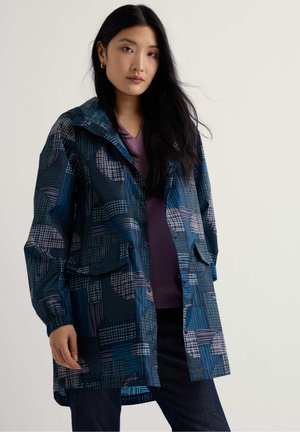 Hosszú sötét hajú nő, aki kék mintás esőkabátot visel lila ing és sötét farmer felett, egyszínű világos háttér előtt áll.