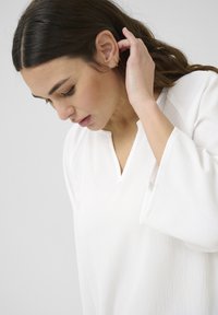 Vit topp med V-ringning, texturerat tyg och vida ärmar. Designen har ett subtilt mönster och bärs av en person som ser ner.
