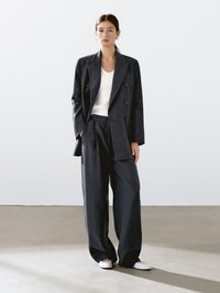 Blazer blu scuro con design doppiopetto, pantaloni a gamba larga coordinati e una t-shirt bianca sotto. Tessuto morbido, accenti minimalisti.