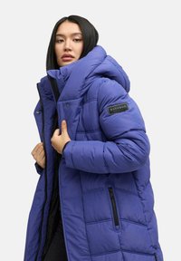 Blauwe pufferjack met een hoge kraag en grote capuchon, voorzien van een doorgestikt patroon, ritszakken aan de zijkant en een logopatch op de mouw.