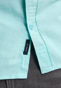 Camicia di lino azzurro chiaro con patta a bottoni, caratterizzata da un'etichetta del marchio blu navy e bordi rifiniti. Il tessuto appare strutturato e traspirante.