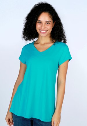 Teal Kurzarm-V-Ausschnitt-T-Shirt mit lockerer Passform. Der Stoff wirkt glatt und leicht. Modell trägt es mit dunkelblauen Jeans.