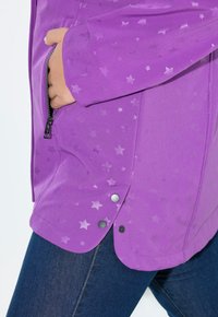 Henkilö, jolla on ykkönen tähtikuviollinen violetti takki, käsi vetoketjulla varustetussa taskussa ja tummansiniset farkut näkyvissä reiteen puolivälistä alaspäin.