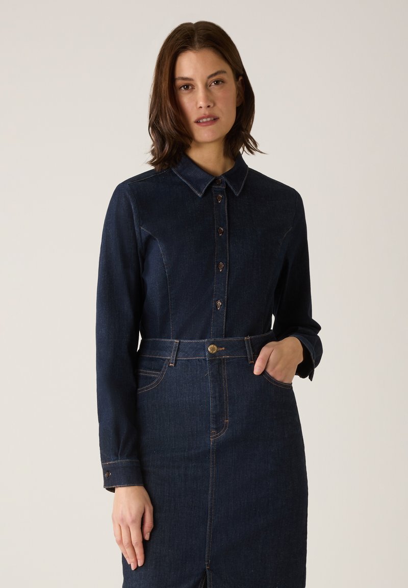 Robe-chemise en denim bleu foncé avec un col à revers, une fermeture boutonnée à l'avant, des manches longues et une taille cintrée. Présente des accents de coutures apparentes.