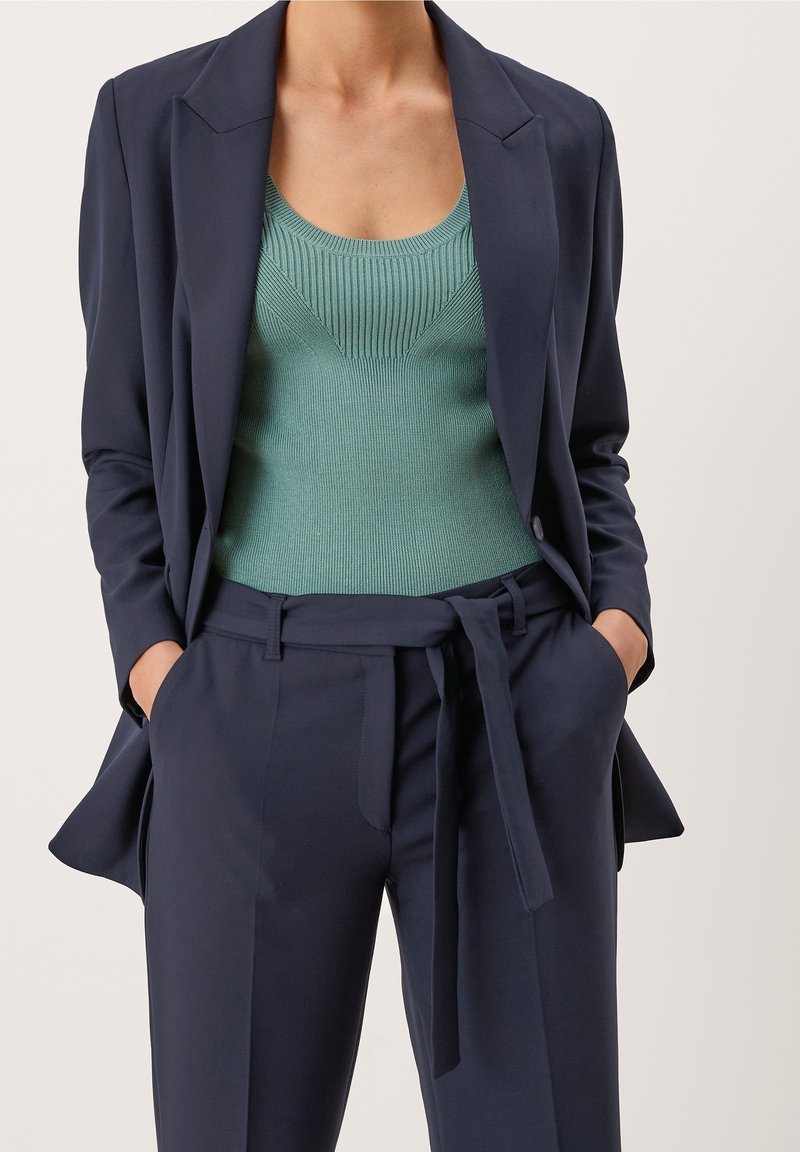 Femme portant un costume bleu marine ajusté avec ceinture en forme de cravate et un top vert côtelé sans manches, mains dans les poches, sur fond neutre.