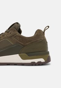 Grüne Wildleder- und Leder-Sneaker mit strukturiertem Finish, ausgestattet mit einer weißen Zwischensohle, einem Zugband und geprägtem Branding an der Ferse.