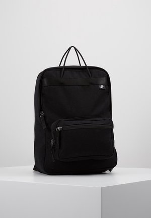 Schwarzer Nike-Rucksack mit Oberhandgriffen, einer vorderen Reißverschlusstasche und minimalistischer Gestaltung, platziert auf einem weißen Podest vor einem schlichten Hintergrund.