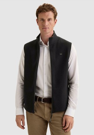 Jeune homme portant un gilet noir sans manches zippé par-dessus une chemise blanche boutonnée et un pantalon beige, debout devant un fond clair uni.