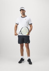 Chemise blanche à manches courtes avec des bordures noires, shorts bleu marine avec le logo Nike, tenant une raquette de tennis, portant des chaussures noires et une visière blanche.