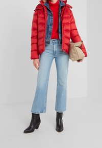Doudoune rouge superposée sur une veste en denim, associée à un jean flare bleu clair et des bottines noires. Tenant un sac beige.