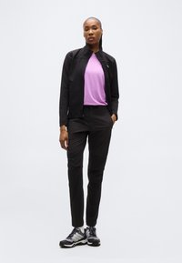 Γυναίκα όρθια, φορώντας μαύρο μπουφάν, λεβαντίνιο μπλουζάκι, μαύρο παντελόνι και μαύρα και άσπρα αθλητικά παπούτσια, μπροστά σε απλό φωτεινό φόντο.