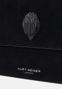 Czarna torba z zamszu z teksturowanym czarnym zapięciem w kształcie liścia. Logo „Kurt Geiger London” w białym kolorze na dole.