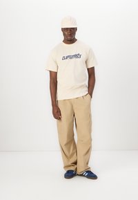 Beige cotton t-shirt with "CLEPTOMANICX" text, paired with beige cargo pants and navy shoes featuring white stripes. Light cap completes the look.