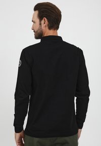 Černé dlouhé rukávy polo tričko vyrobené z bavlny, s límečkem a vyšitým emblémem na levém rukávu. Jednoduché, v jednobarevném provedení bez vzorů.