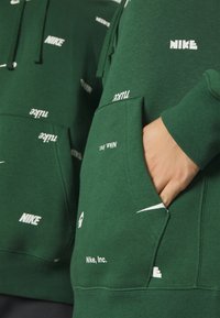 Grön hoodie med flera vita Nike-logotyper, kenguruficka, dragsko i huvan och ribbad mudd. Tillverkad av mjukt, strukturerat tyg.