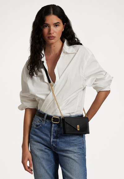 Bolso cruzado de cuero negro con una correa de cadena dorada, que presenta un diseño de solapa angular y un broche frontal. Llevado sobre una camisa blanca y jeans azules.
