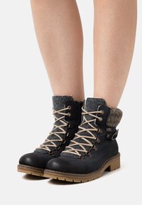 Bottes à lacets noires avec col en feutre gris, lacets beiges et semelles en caoutchouc. Comprend des œillets en métal et un détail de sangle décoratif.