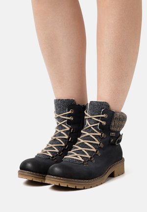 Bottes à lacets noires avec col en feutre gris, lacets beiges et semelles en caoutchouc. Comprend des œillets en métal et un détail de sangle décoratif.