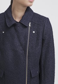 Kobieta nosząca ciemnoniebieską kurtkę o fakturowym wykończeniu z asymetrycznym srebrnym zamkiem i kieszeniami na klapki, zwrócona nieco w lewo.