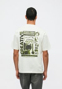 Man i vit t-shirt med abstrakt grön och svart grafik samt text inklusive "KREATIV," "FANTASI," och evenemangsdetaljer på baksidan.