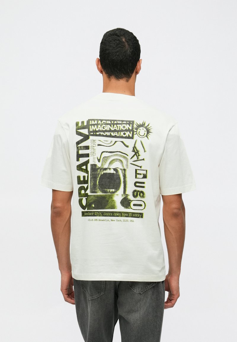 Man i vit t-shirt med abstrakt grön och svart grafik samt text inklusive "KREATIV," "FANTASI," och evenemangsdetaljer på baksidan.