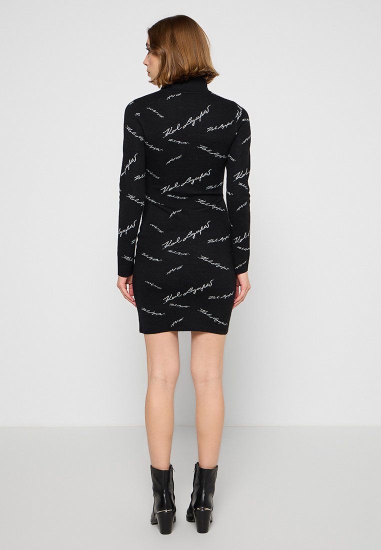 Robe en tricot noir à manches longues et col haut, avec un motif de texte en écriture blanche sur toute la surface. Silhouette ajustée, longueur mini.