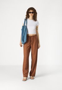 Pantalons en lin marron avec une taille à cordon, associés à un t-shirt blanc ajusté et un sac fourre-tout bleu. Portés avec des sandales beiges.