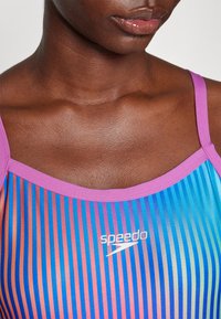 Speedo peldkostīms ar vertikālām gradienta svītrām zilo, rozā un oranžā krāsā. Akcentēts ar gaismas purpura krāsas apkakli un logotipa detaļu.