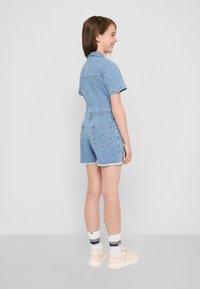 Combinaison en jean courte de couleur bleu clair, avec des manches courtes, un col et des poches au dos. Comprend des bords effilochés et une texture de tissu usé.
