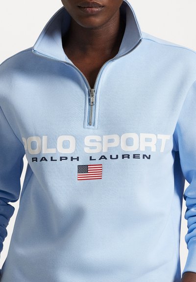 Jasnoniebieski półgolf z zamkiem błyskawicznym wykonany z teksturowanej tkaniny, z białym nadrukiem "POLO SPORT" i "RALPH LAUREN", z małym akcentem w postaci amerykańskiej flagi.