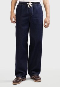Dunkelblaue Denim-Hosen mit entspanntem Schnitt, elastischem Bund, Kordelzugdetail und sichtbaren Nähten. Kombiniert mit braunen Schuhen.